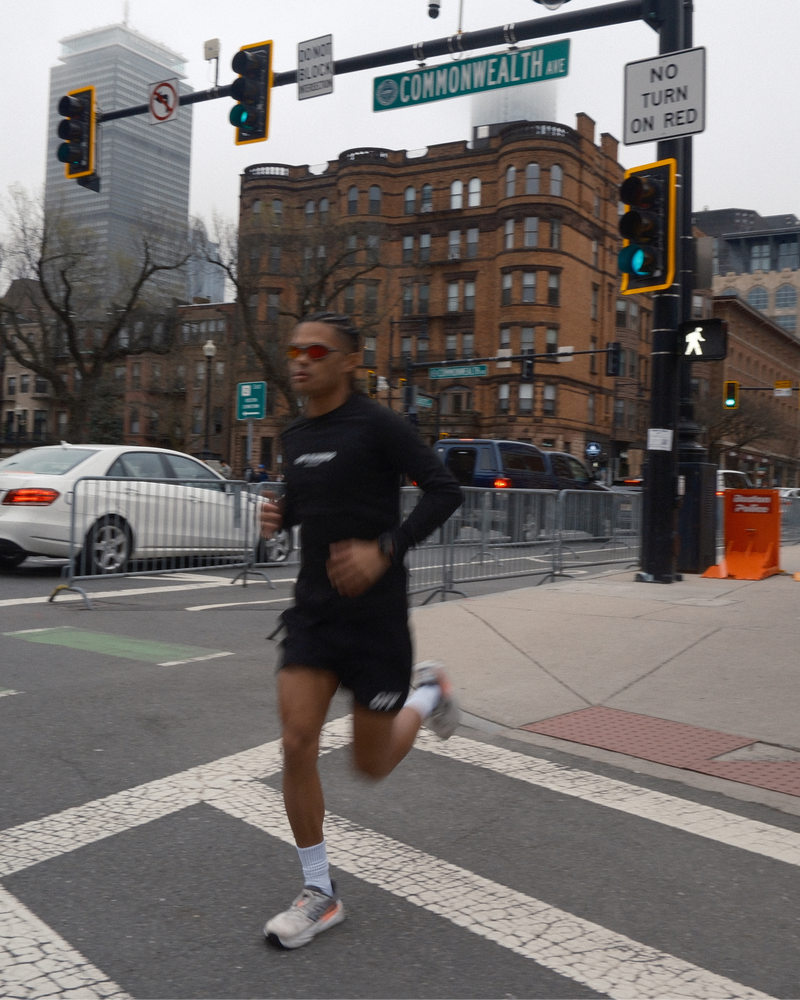 A man wearing a black long-sleeve athletic shirt and black shorts runs across a city crosswalk, with traffic lights, street signs, cars, and brick buildings in the background.